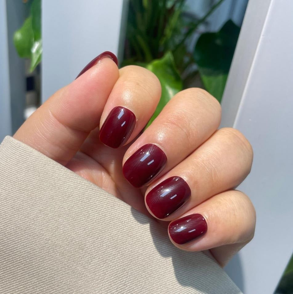 Hand with glossy, short square burgundy press-on nails, styled with a natural light background and subtle silver reflections.