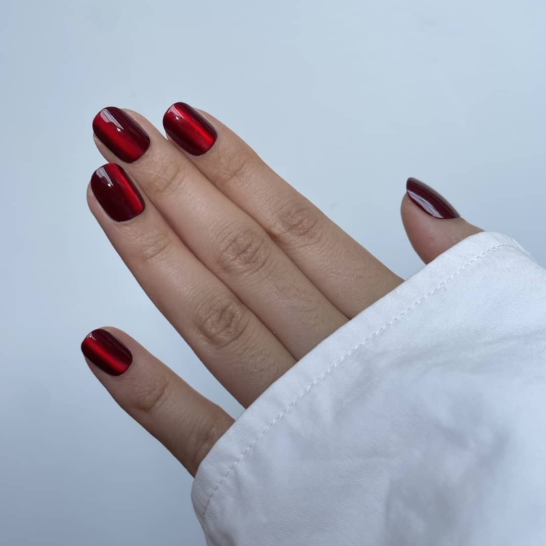 Hand model wearing Ruby Vibe cat-eye magnetic press-on nails in a deep burgundy-red chrome finish on a white background.
